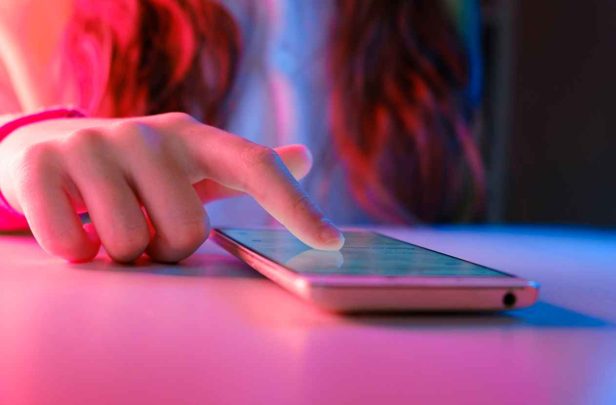 Use of mobile phone in trendy neon lights. Creative vivid color of ultraviolet red and blue. Hands of Teen Girl scrolling up photos Close-up at dark neon room.