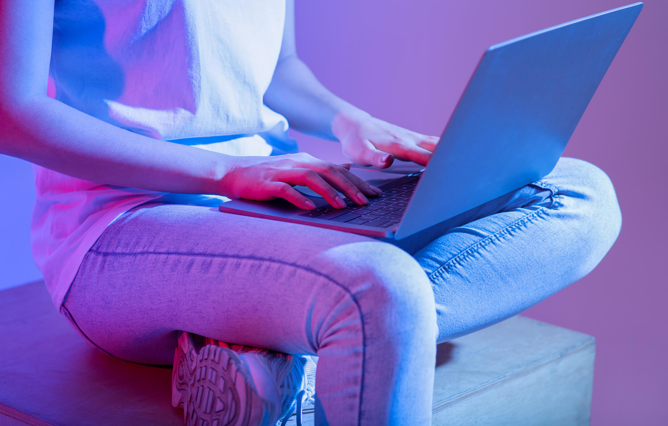 A close up shot of a woman in casual attire sitting on the couch typing on a laptop with a neon purple studio-like background