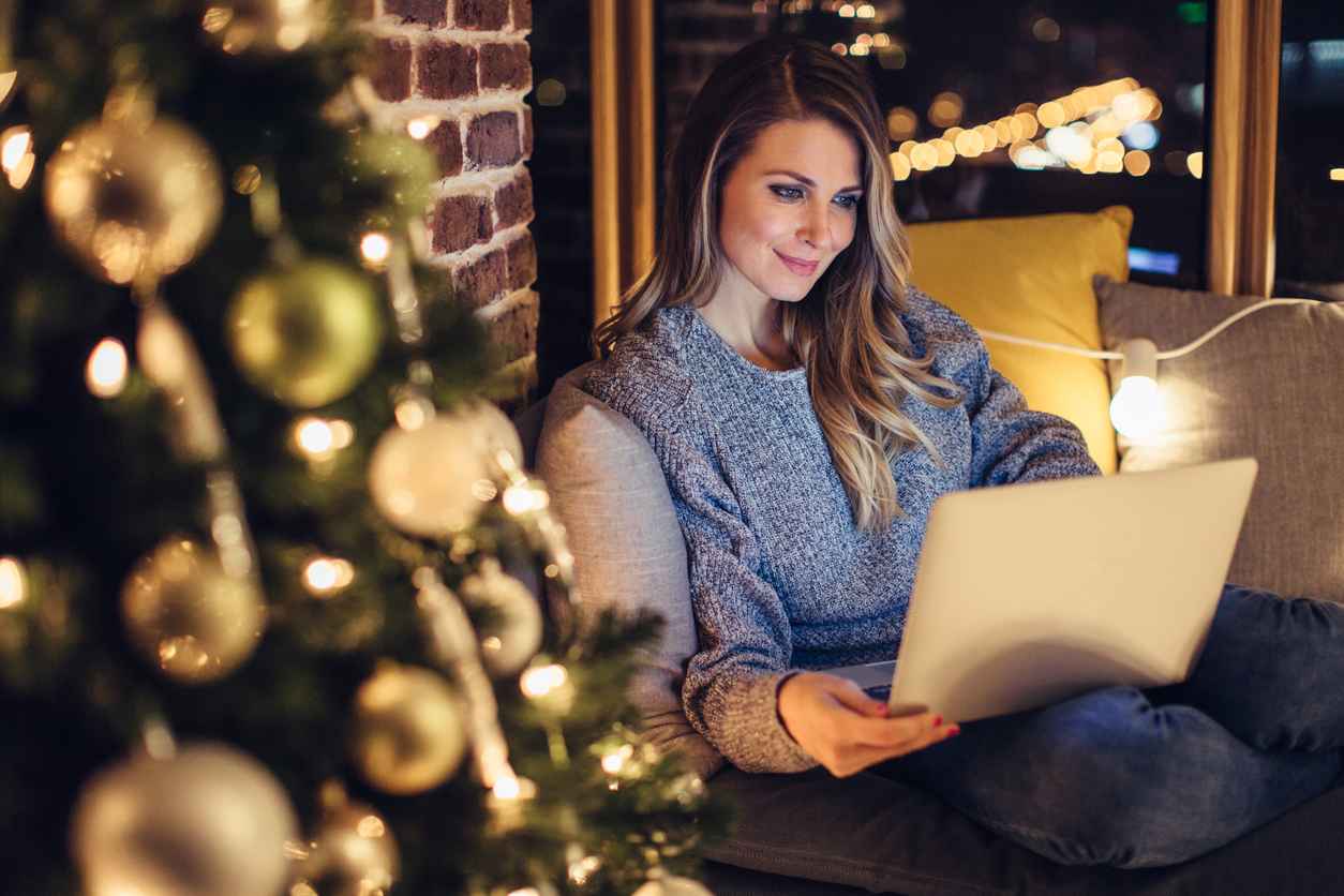 Women on couch next to the Christmas tree, spending beautiful Christmas night at home. She's holding a laptop on her knees.